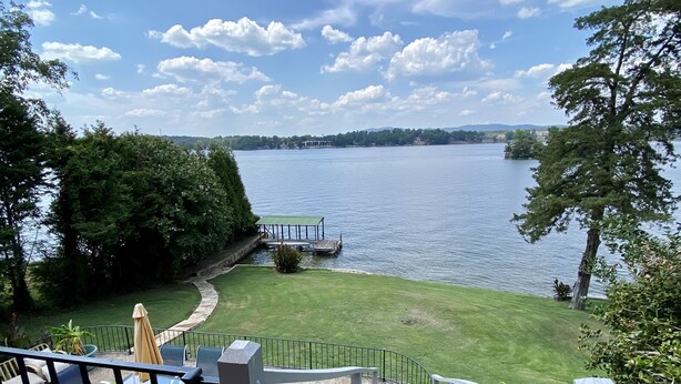 A view of a lake from a balcony with a grassy deck, a dock, and a table with chairs.