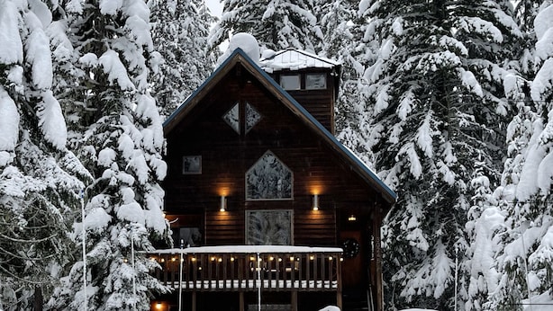 Una cabaña de troncos con fachada de madera, un porche iluminado y árboles cubiertos de nieve.