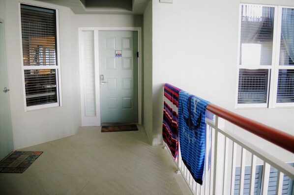 Un couloir avec une porte blanche, une fenêtre avec des stores et une serviette bleue suspendue à une rambarde blanche.