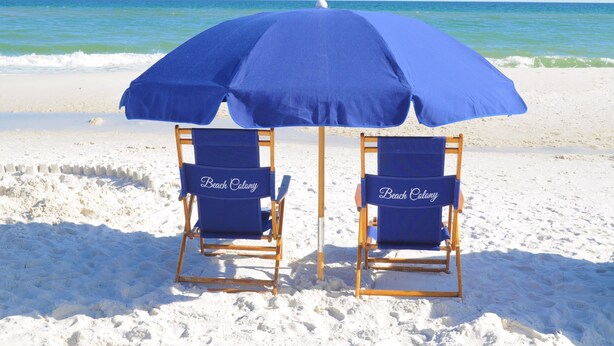 Deux chaises de plage sous un parasol bleu, sur une plage de sable, avec l’océan en arrière-plan.