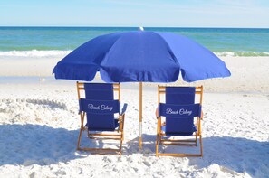 Deux chaises de plage sous un parasol bleu, sur une plage de sable, avec l’océan en arrière-plan.