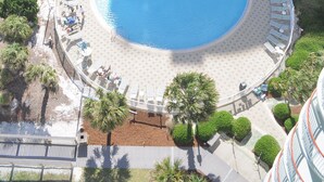 Vue aérienne d’un hôtel doté d’une grande piscine incurvée, entourée de chaises longues et de palmiers.
