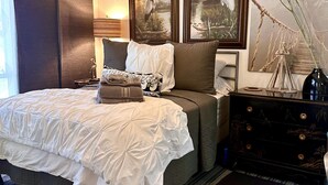 Quarto com cama, mesa de cabeceira, cômoda, ventilador de teto e duas pinturas penduradas na parede.