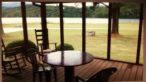 A covered patio with a table and chairs, overlooking a grassy area with a body of water and trees.