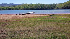 A lakeside scene with a dock extending into the water, surrounded by grassy areas and forested hills in the background.