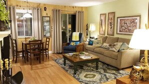 A cozy living room with a wooden floor, a dining table with chairs, a sofa, and a coffee table. There is a window with curtains, a lamp, and framed artwork on the wall. - Everyone Welcome! Private WiFi, Heated Pool, Gas Grills, Catch the View! (Gatlinburg)