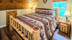 A cozy cabin-style bedroom with a wooden ceiling, a large bed with patterned bedding, a nightstand with a lamp, and a window with a view of trees. - Amazing Cabin w/Firepit, Gametable, Hot Tub, Wifi, Cable (Banner Elk)