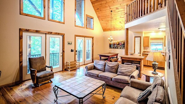 A spacious living room with a high ceiling, wooden beams, and large windows. There is a sofa, a coffee table, and a dining area with a lamp. - Amazing Cabin w/Firepit, Gametable, Hot Tub, Wifi, Cable (Banner Elk)