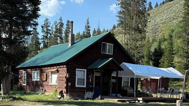 A rustic log cabin with a green metal roof, a porch with outdoor seating, and a covered area with chairs.