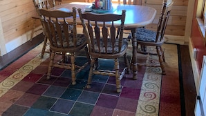 A wooden cabin interior with a round dining table, four wooden chairs, a colorful rug, and a window with lace curtains.