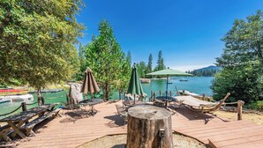 Un muelle de madera con mesas de picnic y sombrillas, con vistas a un lago tranquilo con barcos.