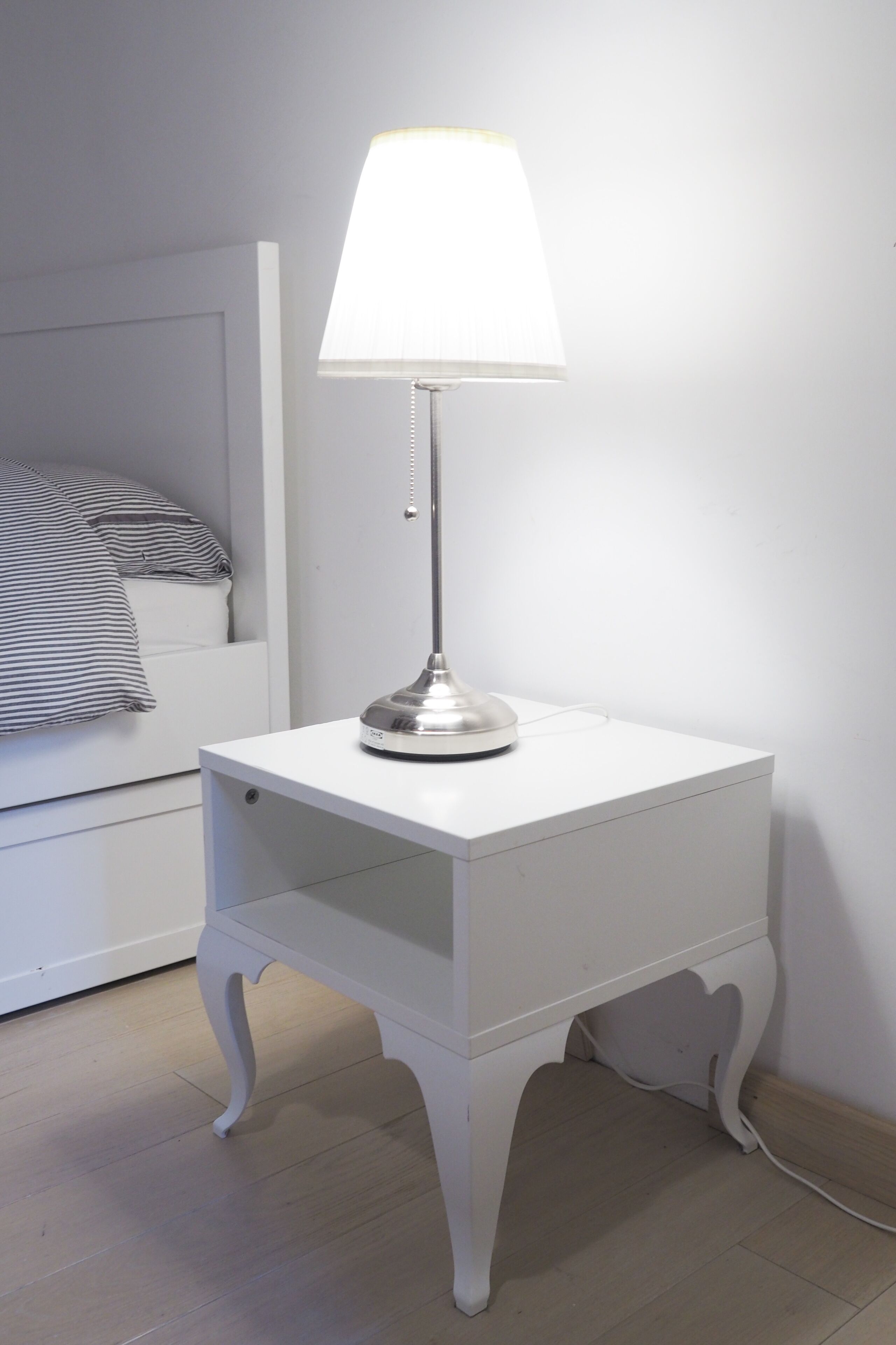 Une table de chevet blanche avec une lampe, un lit avec une literie à rayures et un mur uni.