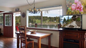 Un coin repas avec une table et des chaises en bois, un lustre, une grande fenêtre donnant sur un lac et une forêt, et un téléviseur diffusant des images de paysages.