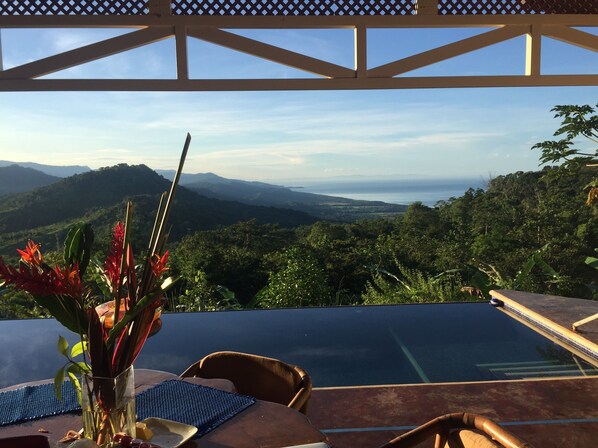 A table with chairs and a vase of flowers, overlooking a pool and a scenic view of mountains and greenery.