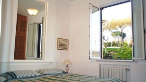 Un dormitorio con cama, una ventana con persiana abierta, una lámpara de noche y un cuadro en la pared.