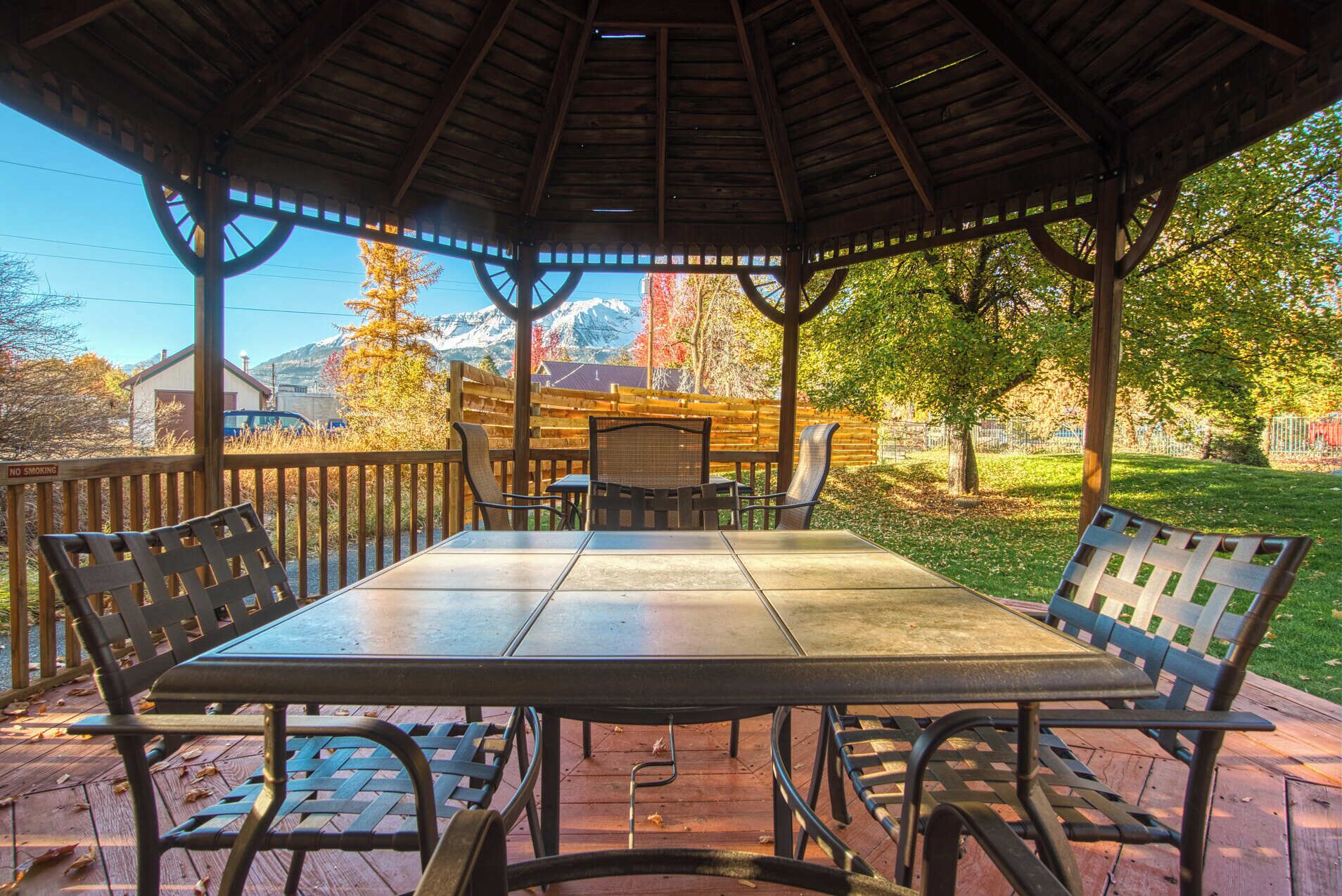A covered outdoor seating area with a table and chairs, surrounded by a wooden fence, overlooking a park with trees and a distant mountain range.
