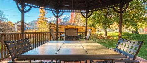 A covered outdoor seating area with a table and chairs, surrounded by a wooden fence, overlooking a park with trees and a distant mountain range.