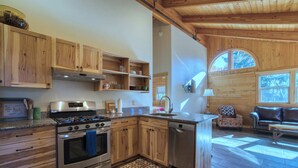 A modern kitchen with wooden cabinets, a stainless steel stove, and a dishwasher. There is a dining area with a patterned rug and a sofa. - Mt. Joseph Lodge in Downtown Joseph: Unit #7 (Joseph)