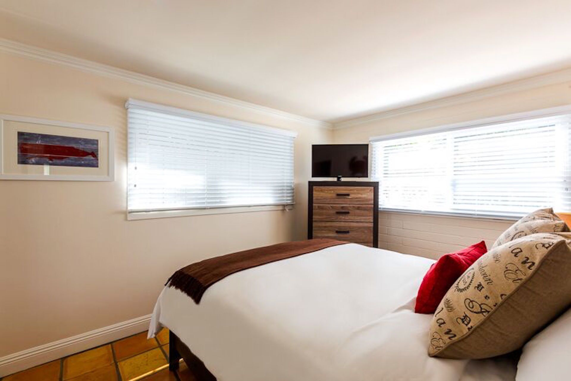 A bedroom with a bed, a dresser, a television, and two windows with blinds.