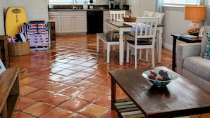 A spacious living area with a dining table, chairs, and a sofa. A kitchen with white cabinets and a window is visible in the background. -  🏄GREAT LOCATION: FAMILY & PET FRIENDLY BEACH COTTAGE🌊 (San Clemente)