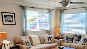A living room with a beige sofa, a wooden coffee table, and a white chair. There is a framed picture on the wall and a window with curtains. -  🏄GREAT LOCATION: FAMILY & PET FRIENDLY BEACH COTTAGE🌊 (San Clemente)