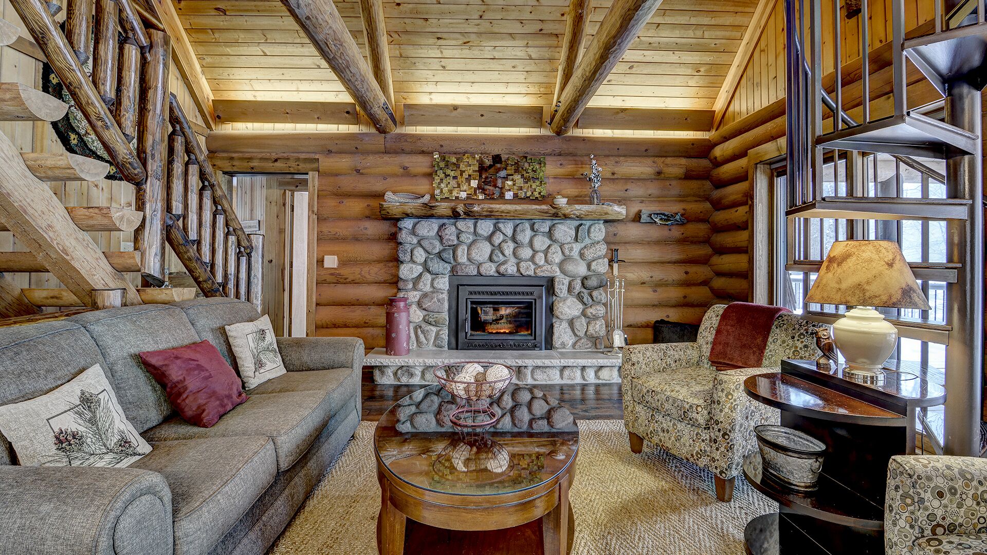 A log cabin interior with a stone fireplace, a sofa, a patterned armchair, and a table with decorative items.