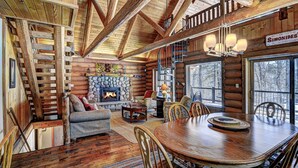 A log cabin interior with a stone fireplace, wooden dining table, and a view of the surrounding forest. - Yellow Birch Lodge - Private Vacation Rental Home       (Eagle River)