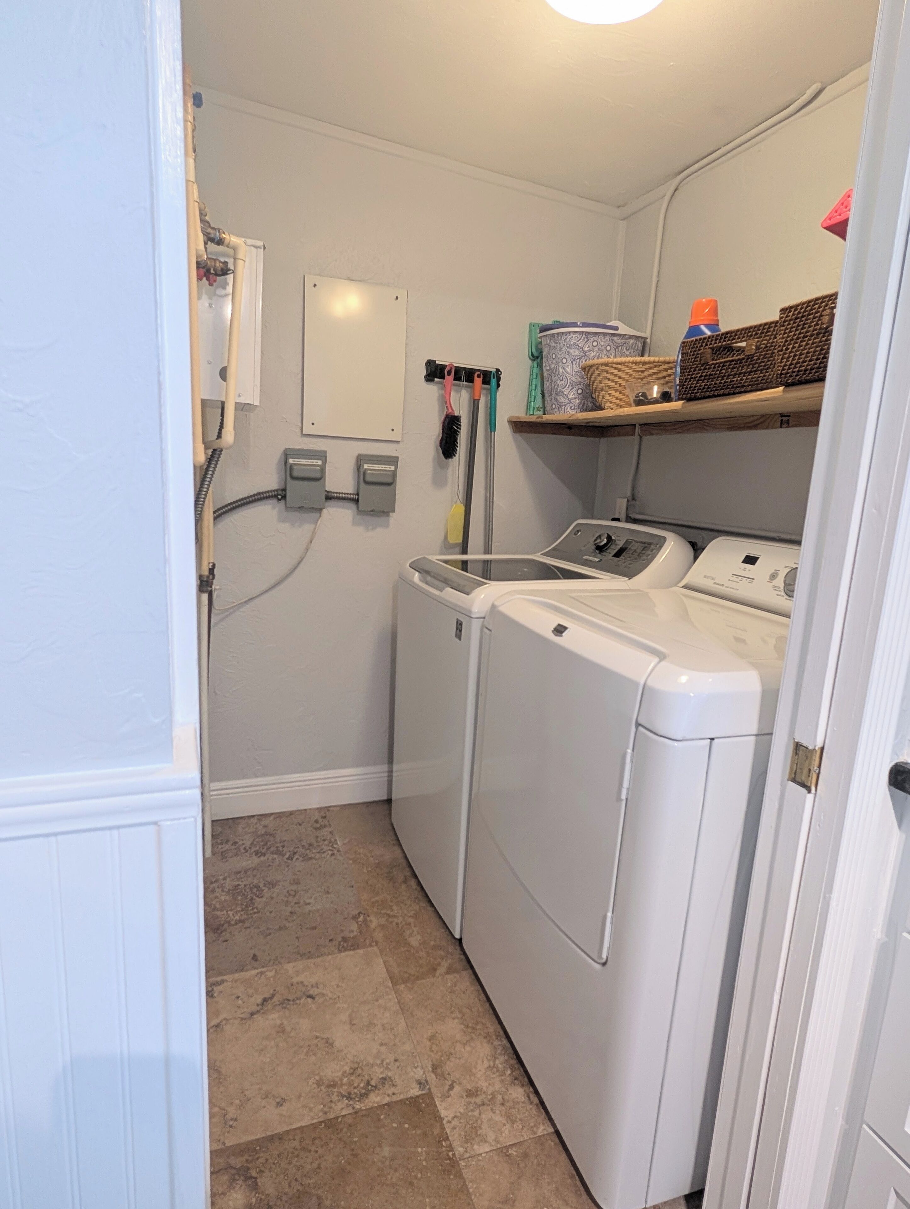 A laundry room with two stacked front-loading washing machines, a drying rack, and a shelf with baskets and a trash can.