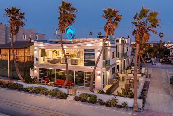 A modern two-story house with a glass facade, outdoor seating, and palm trees.