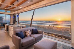 A modern living room with a large window overlooking a beach at sunset, a sofa, and a kitchen area.