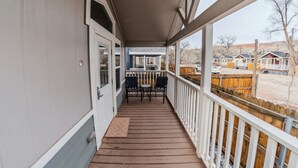 A covered porch with a wooden deck, two chairs, and a view of a fenced area with buildings in the background. - Pet-Friendly Moab Tiny Home Near Arches | Sleeps 6 | Private Porch | Desert View (Moab)