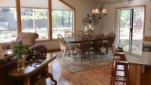 Un espace repas comprenant une grande table, des chaises et un lustre. Il y a une fenêtre avec des stores et une vue sur l’extérieur.