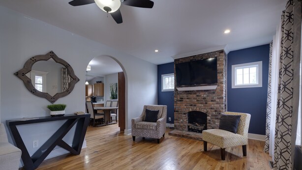 Una sala de estar con piso de madera, chimenea, dos sillones estampados, una consola oscura y un ventilador de techo.