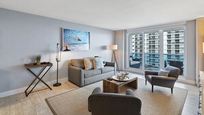 Un salon moderne avec un canapé gris, une table basse et un lampadaire. Une grande fenêtre offre une vue sur les immeubles de grande hauteur.