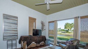 Un salón amplio con un gran ventanal, un ventilador de techo y un televisor de pantalla plana sobre una mesa de madera.