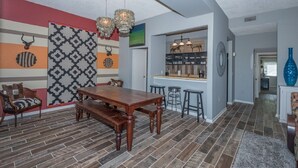 Un comedor con mesa de madera y bancos, pared con diseño y zona de bar con taburetes.
