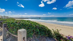 Een strand met een houten omheining, een steiger op de achtergrond en een helderblauwe lucht.