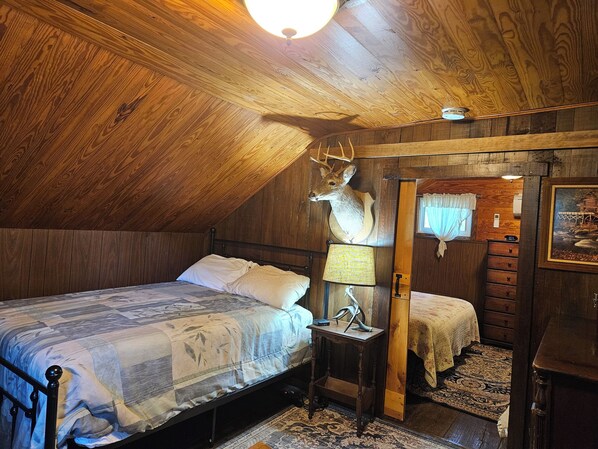 A bedroom with a sloped ceiling, a bed with a patterned bedspread, a nightstand with a lamp, and a deer head mounted on the wall.