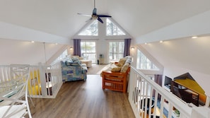 Un loft spacieux avec un plafond haut, un plancher en bois, un coin salon central avec un canapé et des fauteuils, un téléviseur et de grandes fenêtres offrant une vue sur l’extérieur.