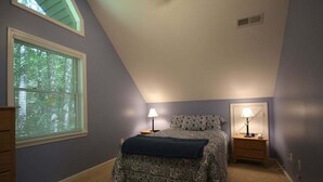 Une chambre avec un plafond en pente, un lit recouvert d’une couvre-lit à motifs, deux tables de chevet avec des lampes et une fenêtre avec des stores.