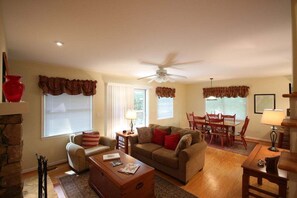 A living room with a fireplace, a ceiling fan, a dining area with a table and chairs, and a large window with curtains.