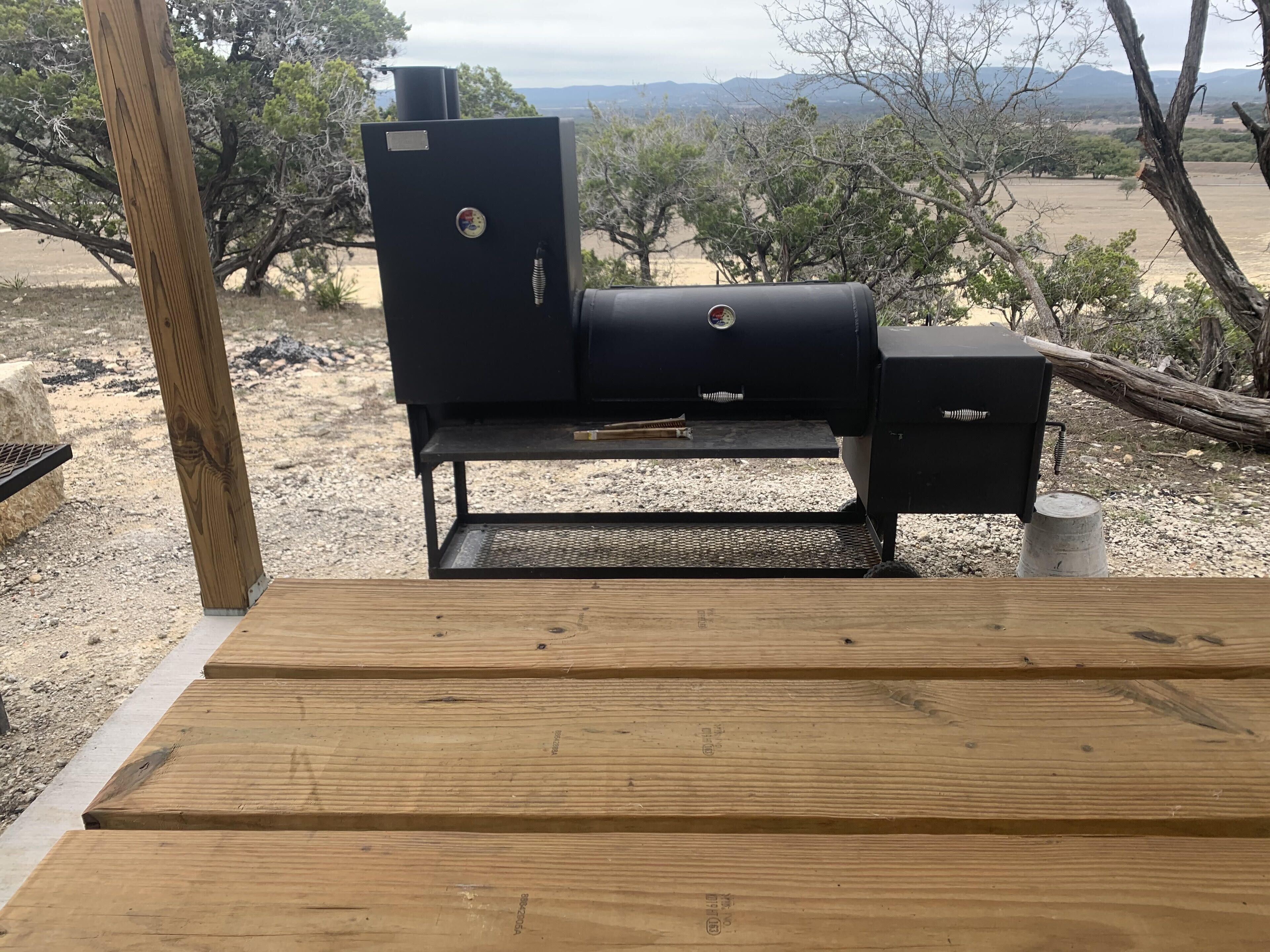 A barbecue grill with a metal grate, a chimney, and a control panel, set on a gravel surface with trees and mountains in the background.