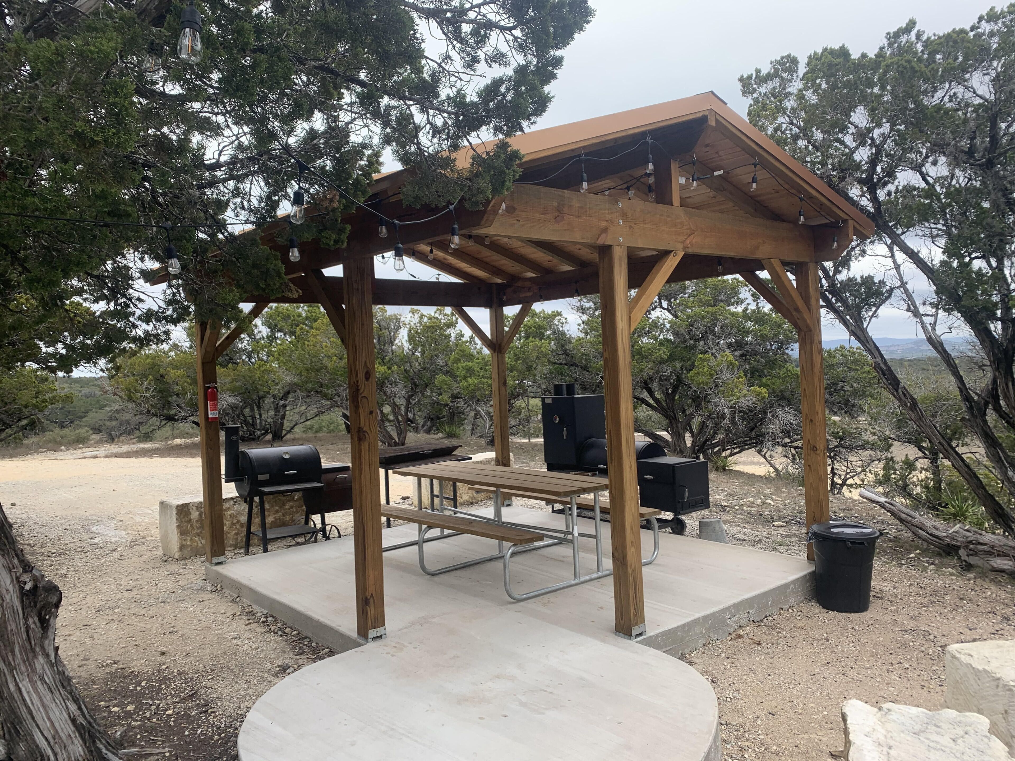 A wooden gazebo with a picnic table and barbecue grills, surrounded by trees and a view of the landscape.