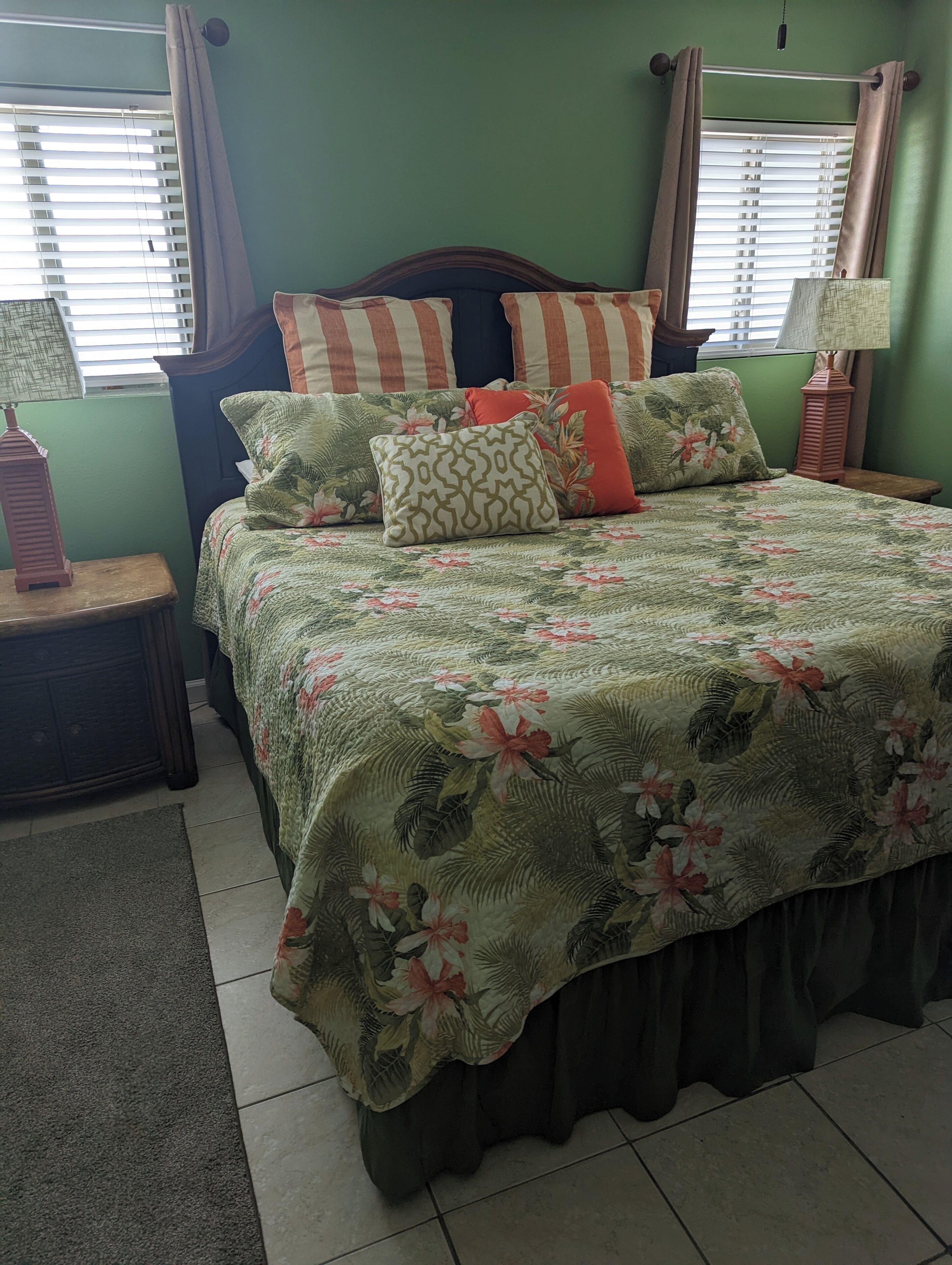 A neatly made bed with floral bedding and striped pillows, a wooden nightstand with a lamp, and a green wall with a window with blinds.