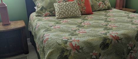 A neatly made bed with floral bedding and striped pillows, a wooden nightstand with a lamp, and a green wall with a window with blinds.