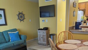 Una sala de estar con un sofá color turquesa, una mesa de centro, una mesa de comedor con sillas, un televisor montado en la pared y una zona de cocina con armarios y un microondas.