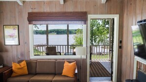 A cozy living room with a brown sofa, orange cushions, a wooden coffee table, and a television. A large window offers a view of a lake and deck area.