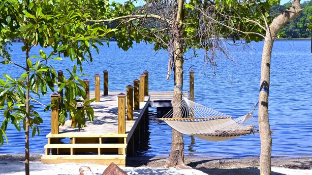 A wooden dock extending into a lake, with a hammock hanging from a tree and a wooden platform with railings.