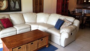 Een woonkamer met een beige hoekbank, een houten salontafel, rieten manden, een blauw vloerkleed, een eethoek met een tafel en stoelen, en een kroonluchter.