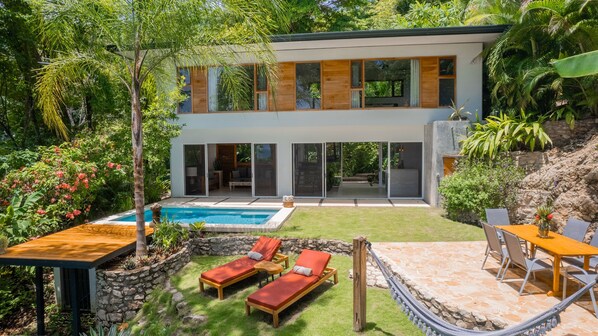 Una casa moderna de dos pisos con piscina, mobiliario exterior y exuberante follaje.